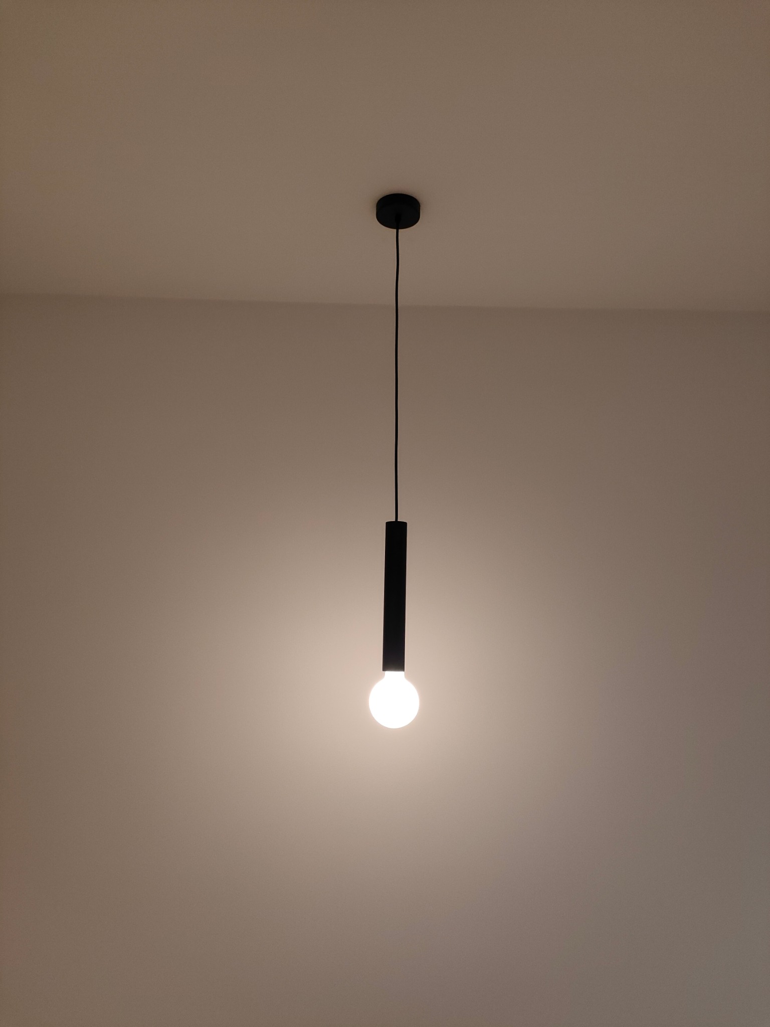 Minimalistyczna czarna lampa wisząca z okrągłą żarówką, zamontowana na białym suficie, oświetlająca beżowe ściany.