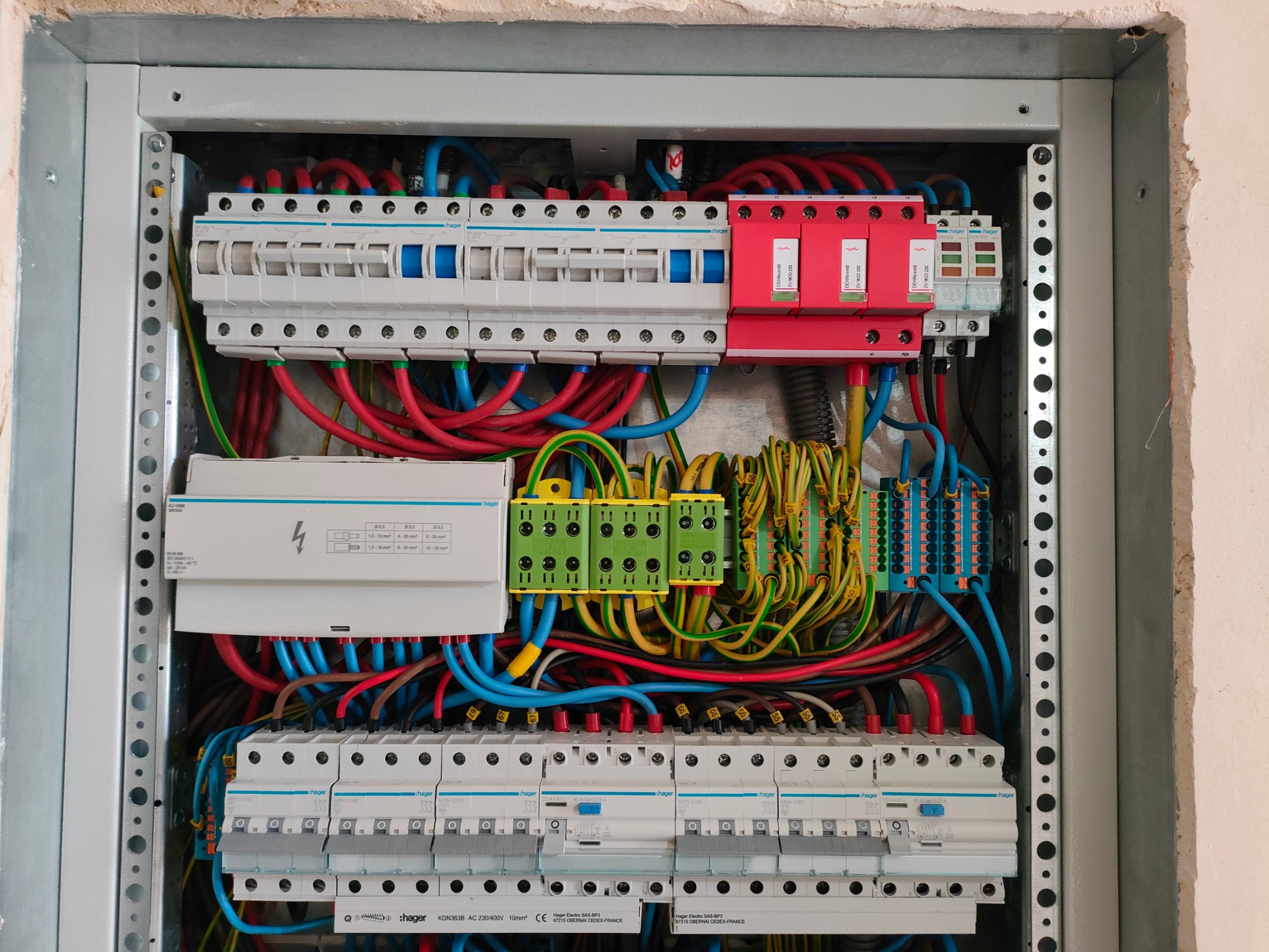 Otwarta skrzynka elektryczna z widocznymi bezpiecznikami, przewodami w różnych kolorach (czerwonym, niebieskim, żółtym, zielonym), listwami zaciskowymi i elementami ochronnymi, zamontowana...