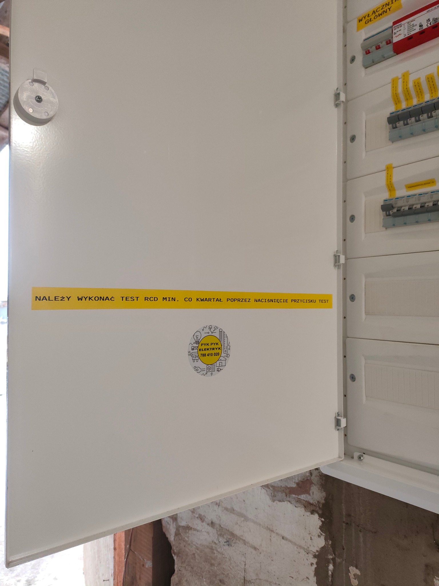 Otwarta skrzynka elektryczna z naklejką informującą o konieczności testowania RCD, widoczne zabezpieczenia i wyłączniki, surowy mur w tle.