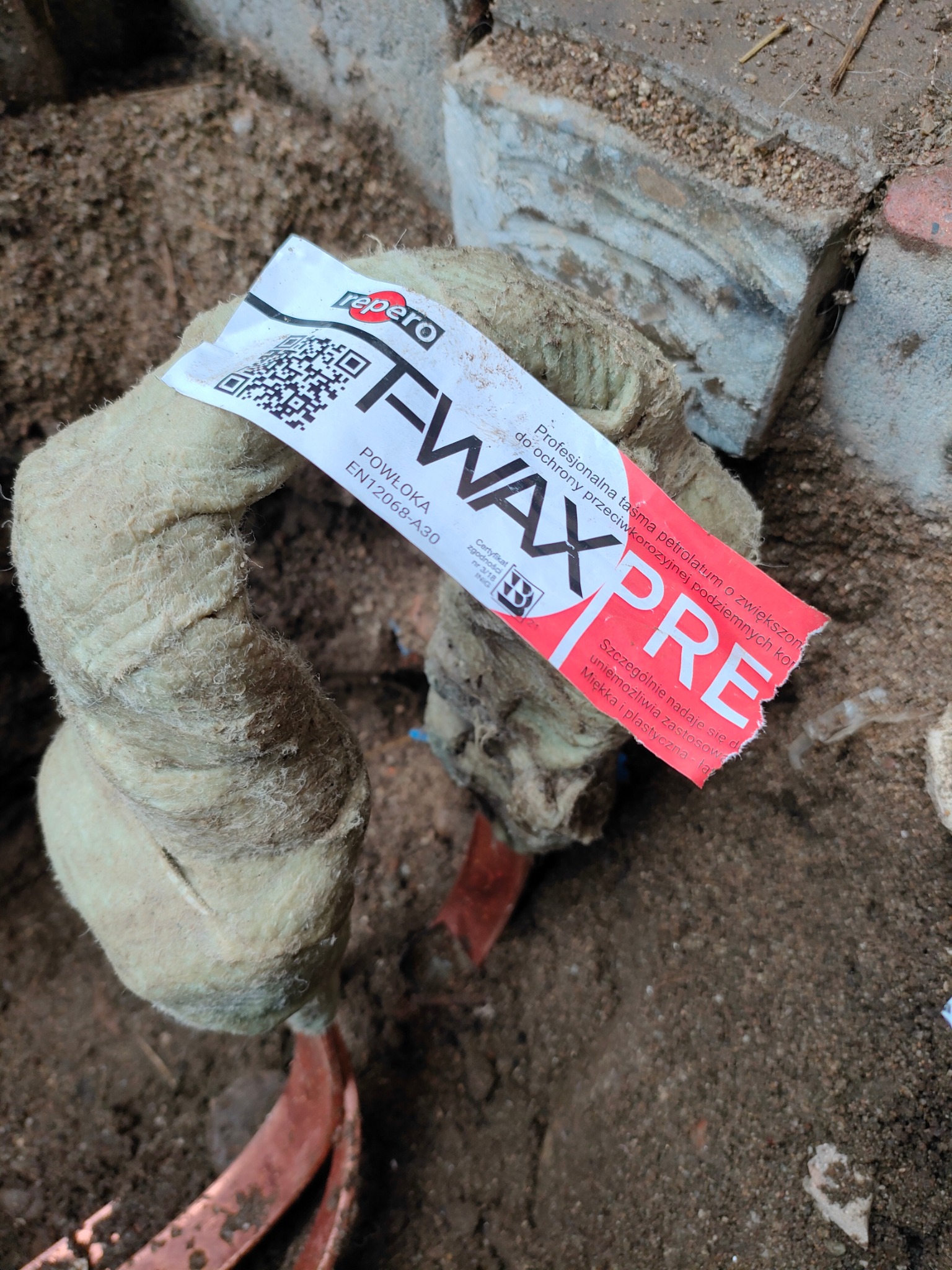 Rura owinięta izolacją z etykietą Repero FWAX PRE, widoczne elementy instalacji w ziemi.