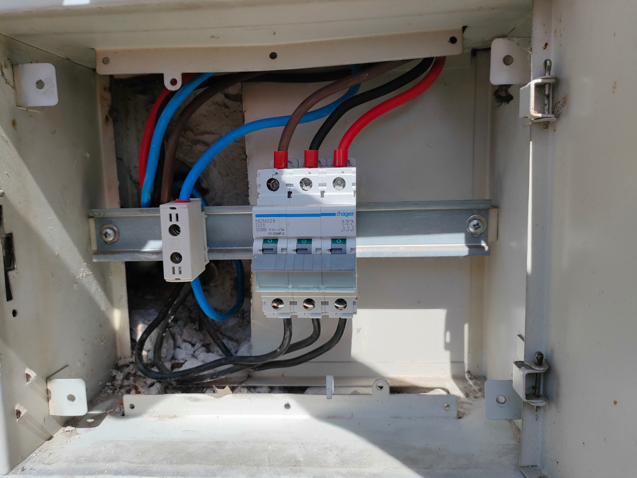 Otwarta skrzynka elektryczna z zamontowanym wyłącznikiem nadprądowym Hager, widoczne przewody w kolorach: niebieskim, brązowym, czarnym i czerwonym, zamocowane na szynie DIN, w tle fragment ściany.
