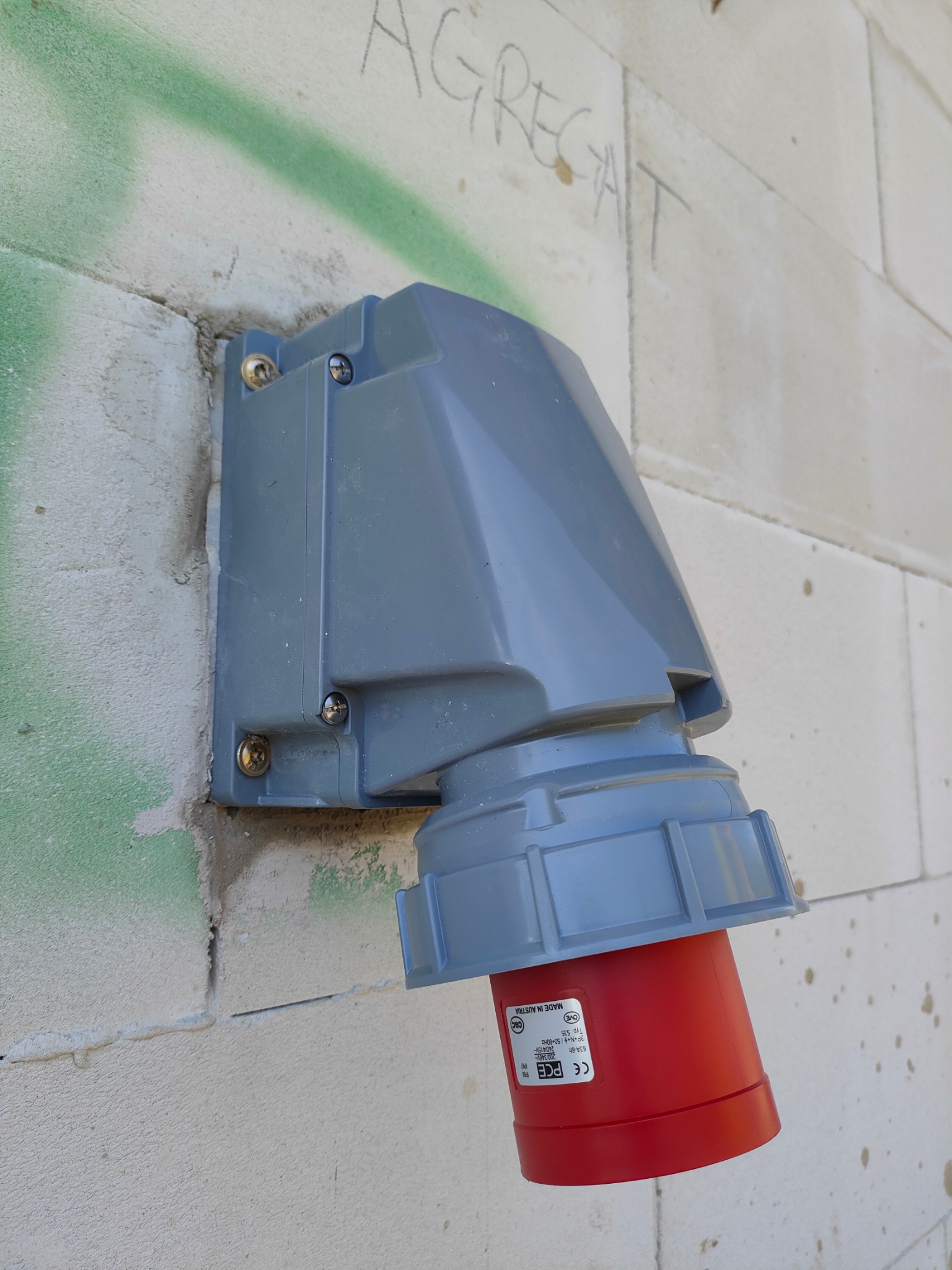 Szara obudowa gniazda elektrycznego przemysłowego z czerwoną wtyczką zamontowana na ścianie z betonu komórkowego z widocznymi śladami zaprawy i napisem 'AGREGAT' wykonanym ołówkiem.