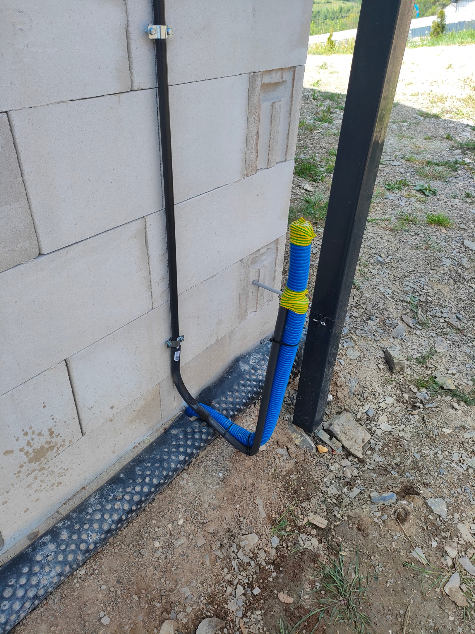 Czarna rura przymocowana do ściany z betonu komórkowego, przechodząca w niebieską rurę karbowaną z żółto-zielonymi oznaczeniami, prowadzącą w ziemię obok czarnego słupka.