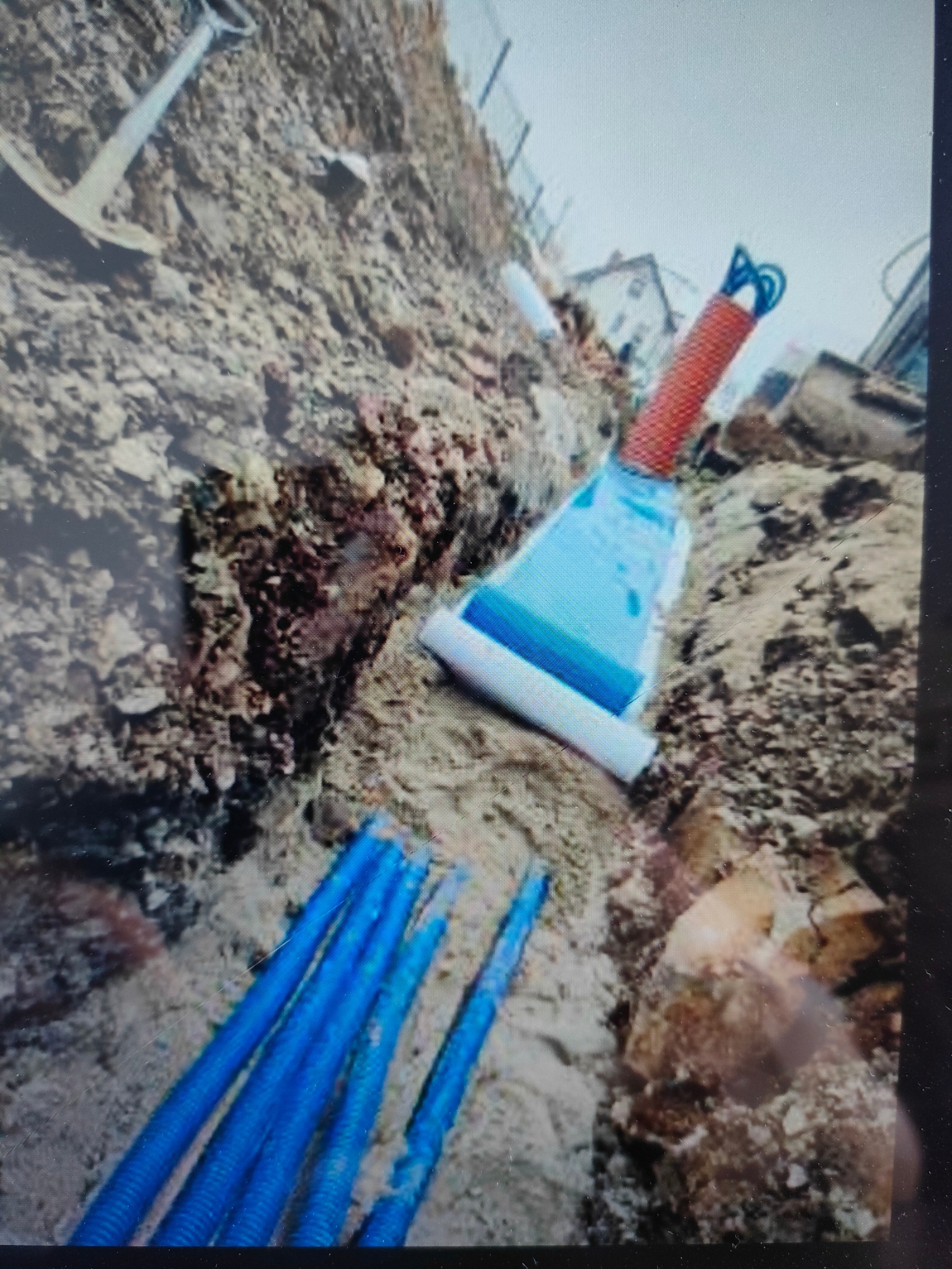 Wykop z ułożonymi rurami osłonowymi kabli elektrycznych, zabezpieczonymi niebieską folią i pomarańczową rurą karbowaną, widoczny kilof wbity w ziemię