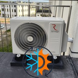 ACTIV KRZYSZTOF LESZCZYŃSKI - Biały agregat klimatyzacji Rotenso Hiro na balkonie, widok z bliska. Częściowo widoczne barierki balkonu i budynki w tle. Nałożony znak graficzny chłodzenia i ogrzewania.