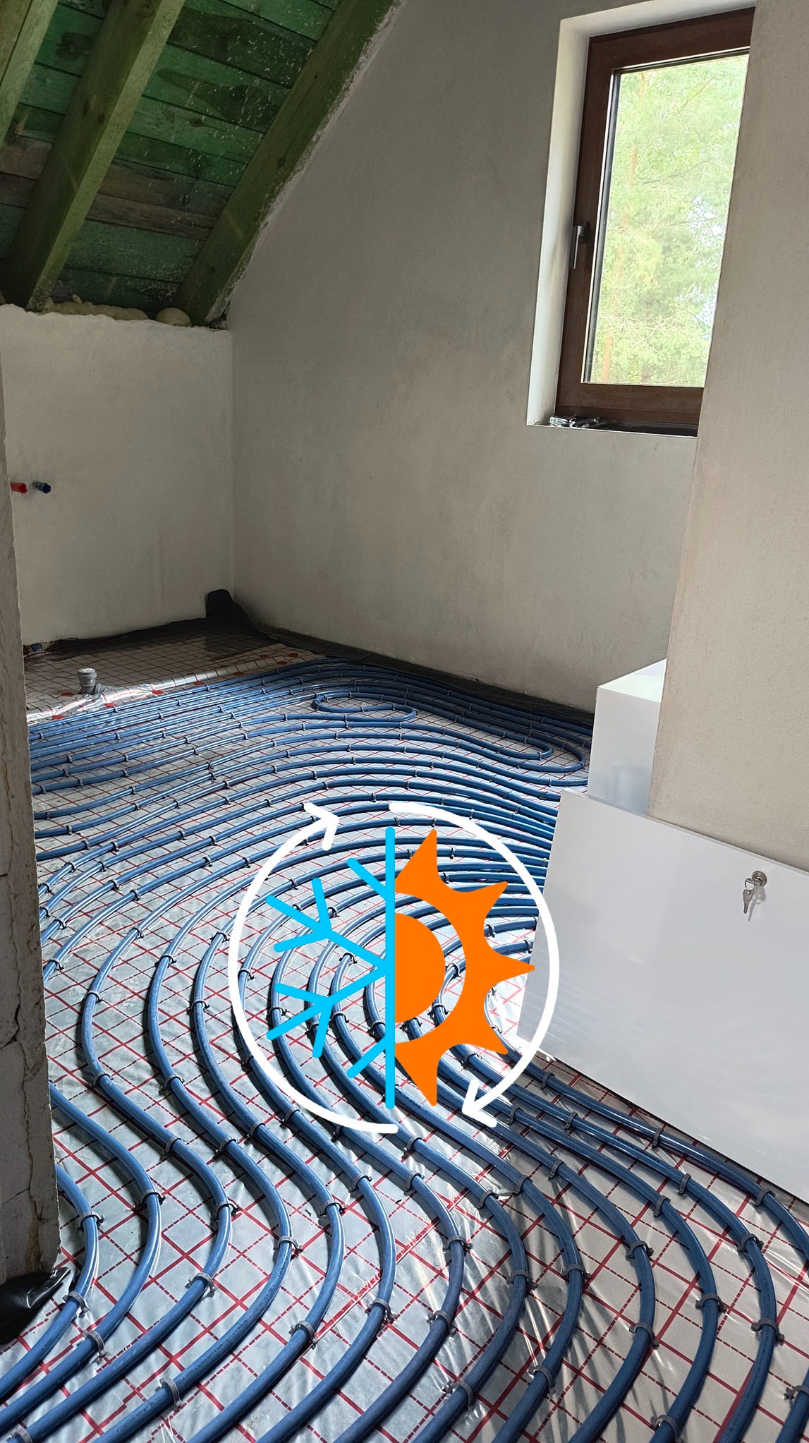 Niebieskie rury ogrzewania podłogowego ułożone spiralnie na folii izolacyjnej w surowym wnętrzu poddasza z oknem. Widoczny fragment zielonej więźby dachowej.