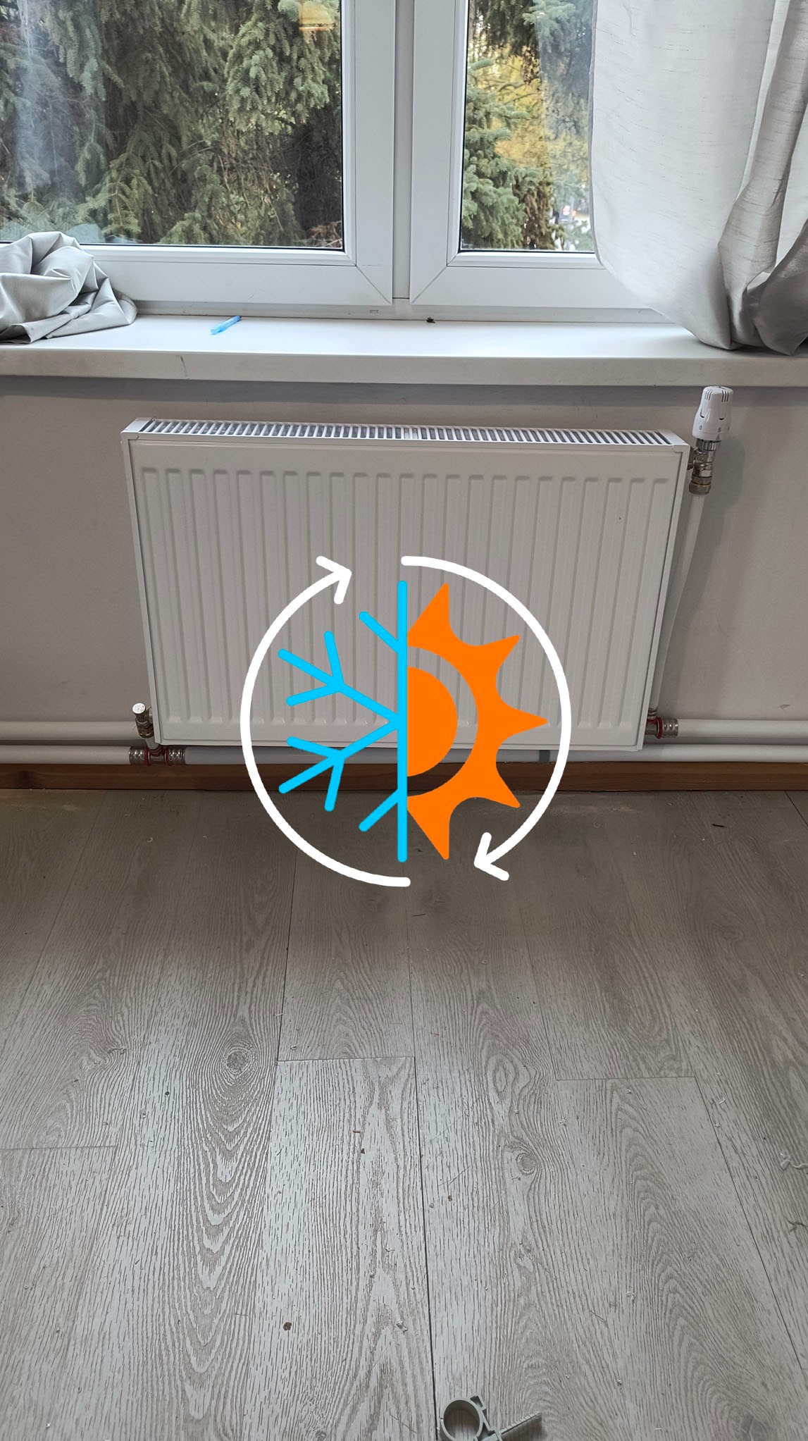 Biały grzejnik panelowy zamontowany pod oknem, z termostatem i rurami doprowadzającymi. Nałożona grafika słońca i śnieżynki w okręgu.