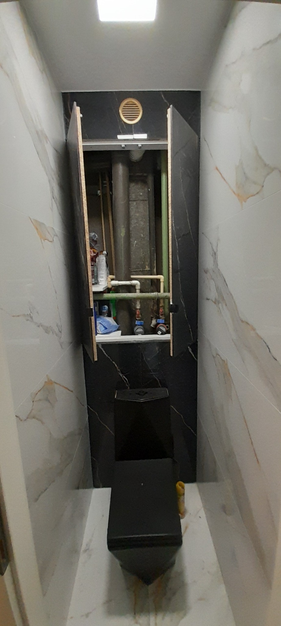 Czarny, wiszący sedes w łazience z marmurowymi płytkami, z otwartą szafką rewizyjną, ukazującą rury i zawory instalacji wodnej.