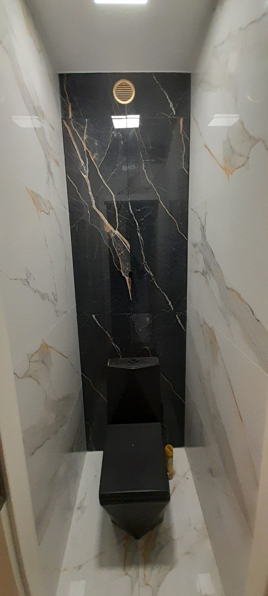 Nowoczesna toaleta w wąskiej łazience, z czarną miską WC i płytkami imitującymi marmur w kolorach czerni i bieli, okrągła kratka wentylacyjna na ścianie.