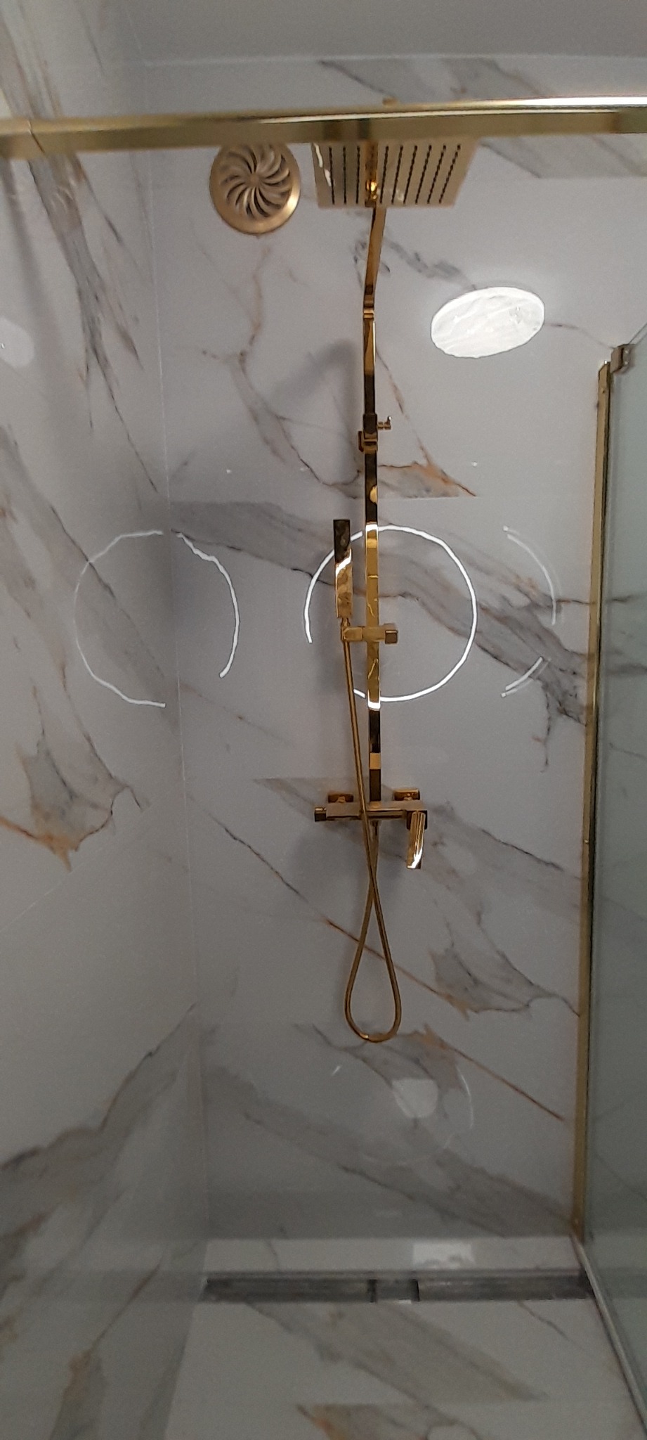 Błyszcząca, złota armatura natryskowa zamontowana na ścianie z marmurowymi płytkami w kabinie prysznicowej, widoczny odpływ liniowy