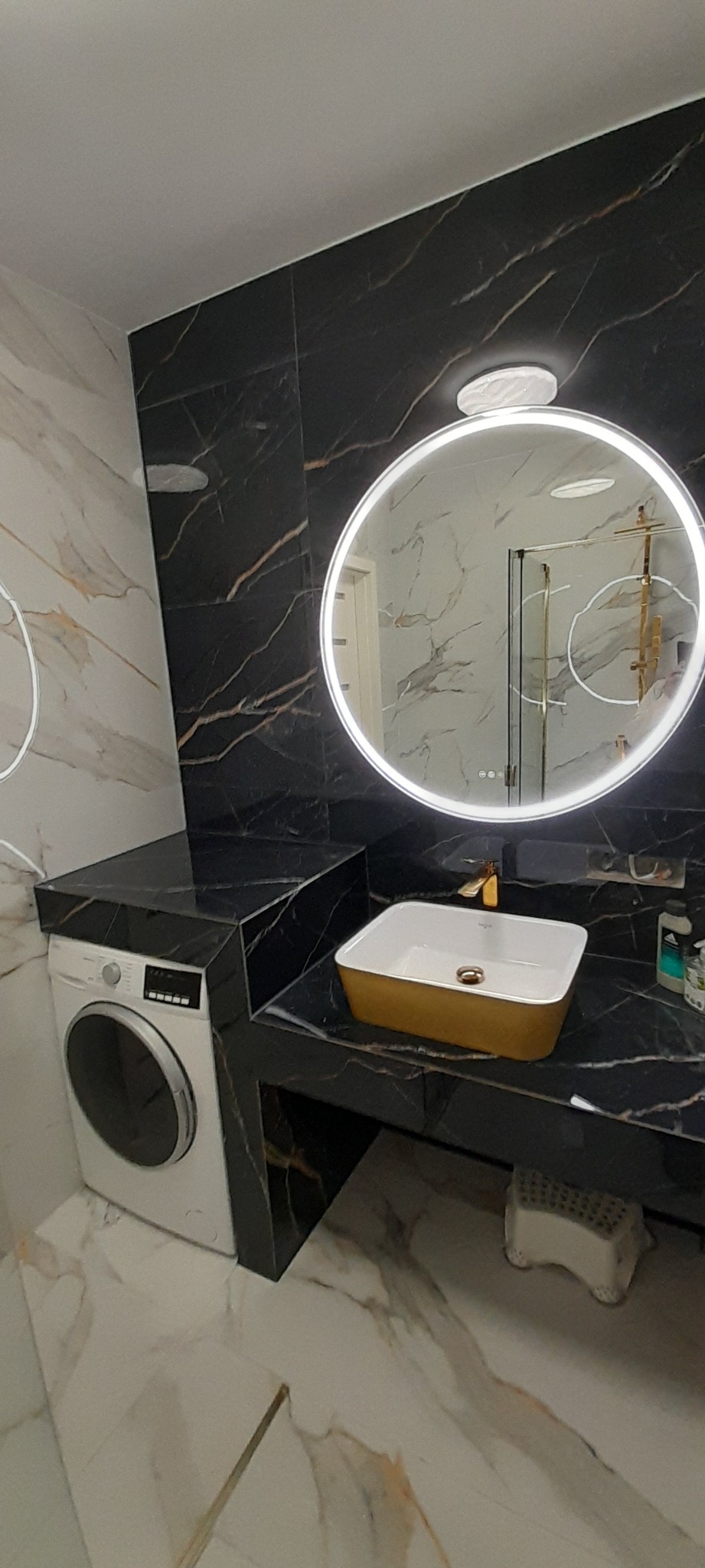 Nowoczesna łazienka z czarnymi marmurowymi płytkami, okrągłym lustrem z podświetleniem LED, złotą umywalką nablatową i pralką w zabudowie pod blatem.