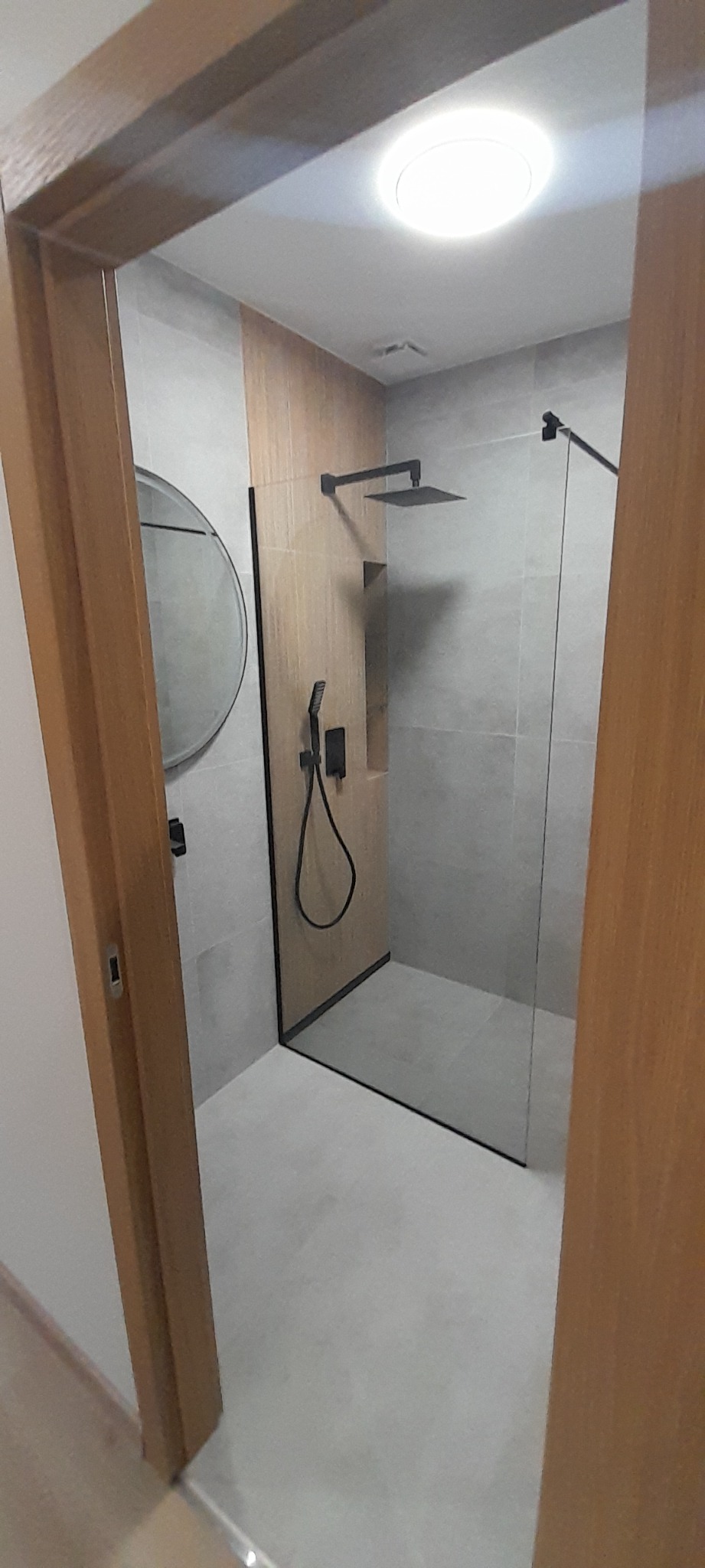 Nowoczesna kabina prysznicowa z czarną armaturą i szklaną ścianką, widoczna przez drewnianą futrynę drzwi, z okrągłym lustrem na ścianie.