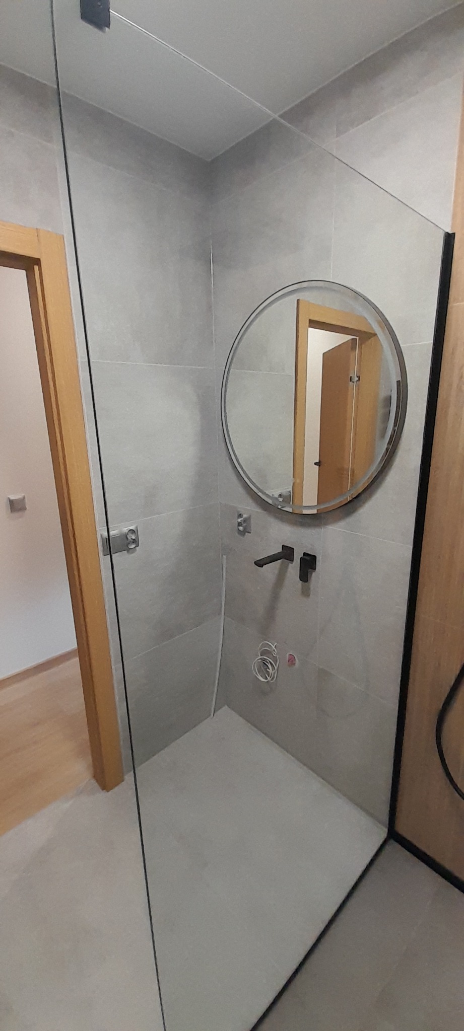 Nowoczesna łazienka z szarymi płytkami, okrągłym lustrem i czarną armaturą, widoczne przygotowane przyłącza wodne, częściowo przeszklona kabina prysznicowa.