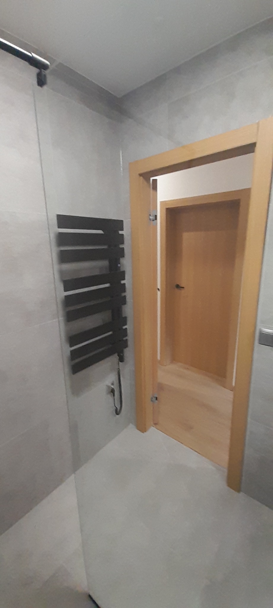 Nowoczesna łazienka z szarymi płytkami, grzejnikiem drabinkowym w kolorze antracytu i otwartymi drzwiami do kolejnego pomieszczenia.