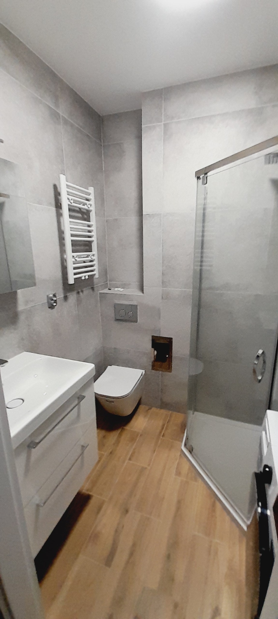 Nowoczesna łazienka po remoncie z szarymi płytkami ściennymi, drewnopodobną podłogą, białą umywalką z szafką, wiszącą toaletą i kabiną prysznicową ze szklanymi drzwiami.