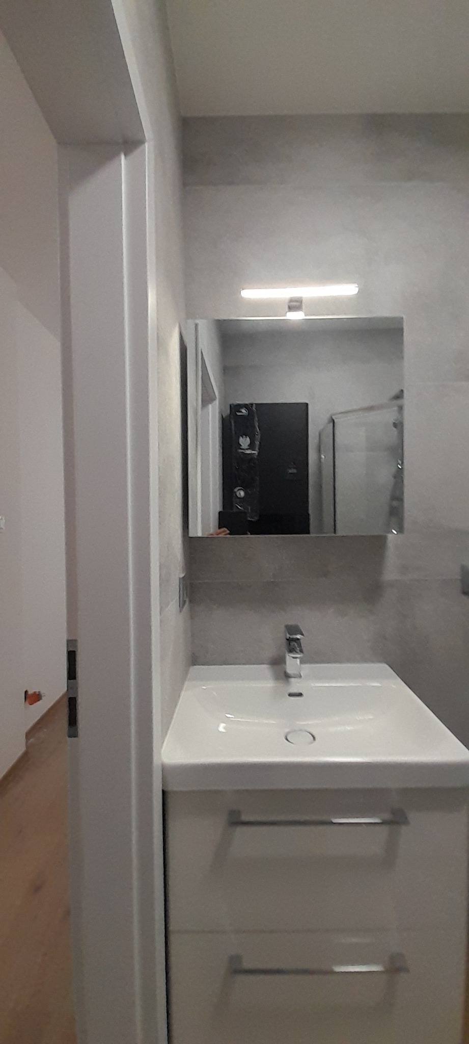 Nowoczesna łazienka z umywalką nablatową na szafce z dwiema szufladami, lustro z oświetleniem LED i szare płytki na ścianie. W odbiciu lustrzanym widoczny czarny regał lub szafka.