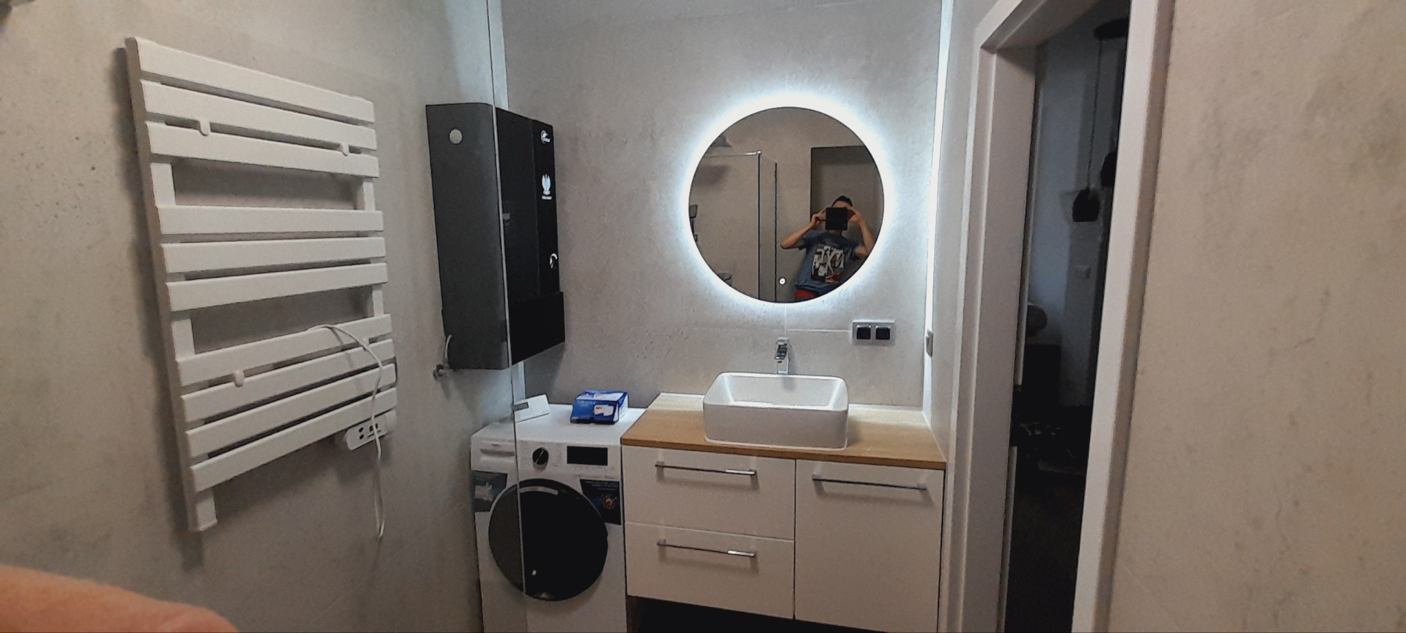 Nowoczesna łazienka z okrągłym podświetlanym lustrem, białą umywalką nablatową na drewnianym blacie, grzejnikiem drabinkowym i czarnym dozownikiem wody nad pralką.