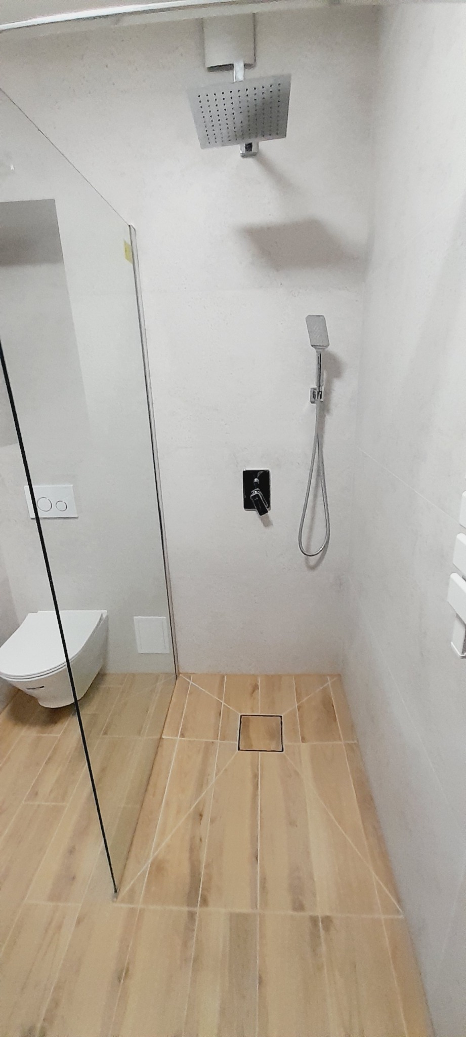 Nowoczesna łazienka z kabiną prysznicową typu walk-in, beżowymi płytkami imitującymi drewno na podłodze, minimalistyczną armaturą w kolorze czarnym i podtynkową spłuczką WC.