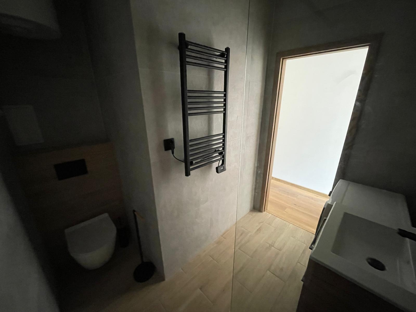 Nowoczesna łazienka z szarymi płytkami, czarnym grzejnikiem drabinkowym elektrycznym, wiszącą toaletą oraz drewnopodobną podłogą i widocznym fragmentem umywalki z szafką.