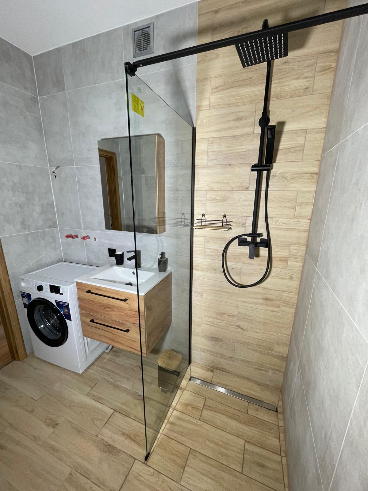 Nowoczesna łazienka z kabiną prysznicową typu walk-in, umywalką nablatową z szafką z drewnianymi frontami, pralką i płytkami imitującymi drewno na podłodze i części ścian.