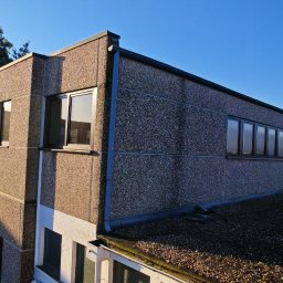 EKOFENSTER.EU SPÓŁKA Z OGRANICZONĄ ODPOWIEDZIALNOŚCIĄ - Widok z góry na budynek z oknami w czarnych ramach, elewacja z szarego betonu architektonicznego, na dachu widoczny agregat klimatyzacji i drabina.