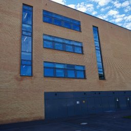 EKOFENSTER.EU SPÓŁKA Z OGRANICZONĄ ODPOWIEDZIALNOŚCIĄ - Fasada budynku z cegły klinkierowej z niebieskimi oknami i szarymi drzwiami na parterze, błękitne niebo z obłokami w tle.