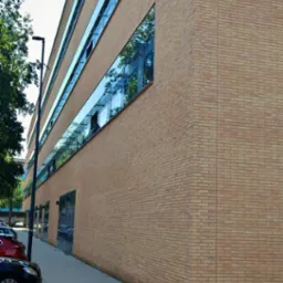 Nowoczesny budynek biurowy z elewacją z jasnej cegły i pasmowymi oknami z ciemnymi ramami, odbijającymi zieleń drzew i błękit nieba.