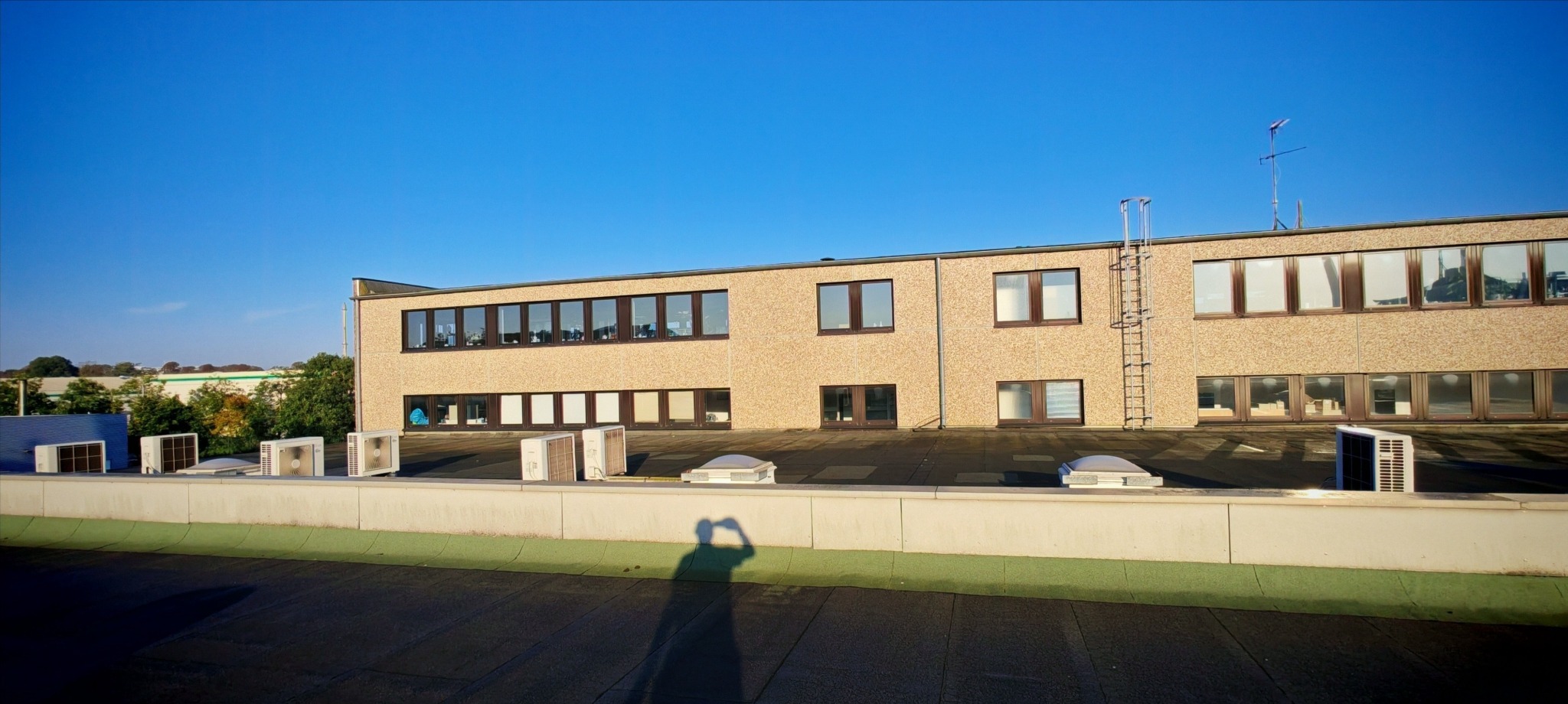 Widok z dachu budynku w Somoninie na fasadę z oknami, klimatyzatory i świetliki dachowe. Na dachu widoczny cień fotografa.