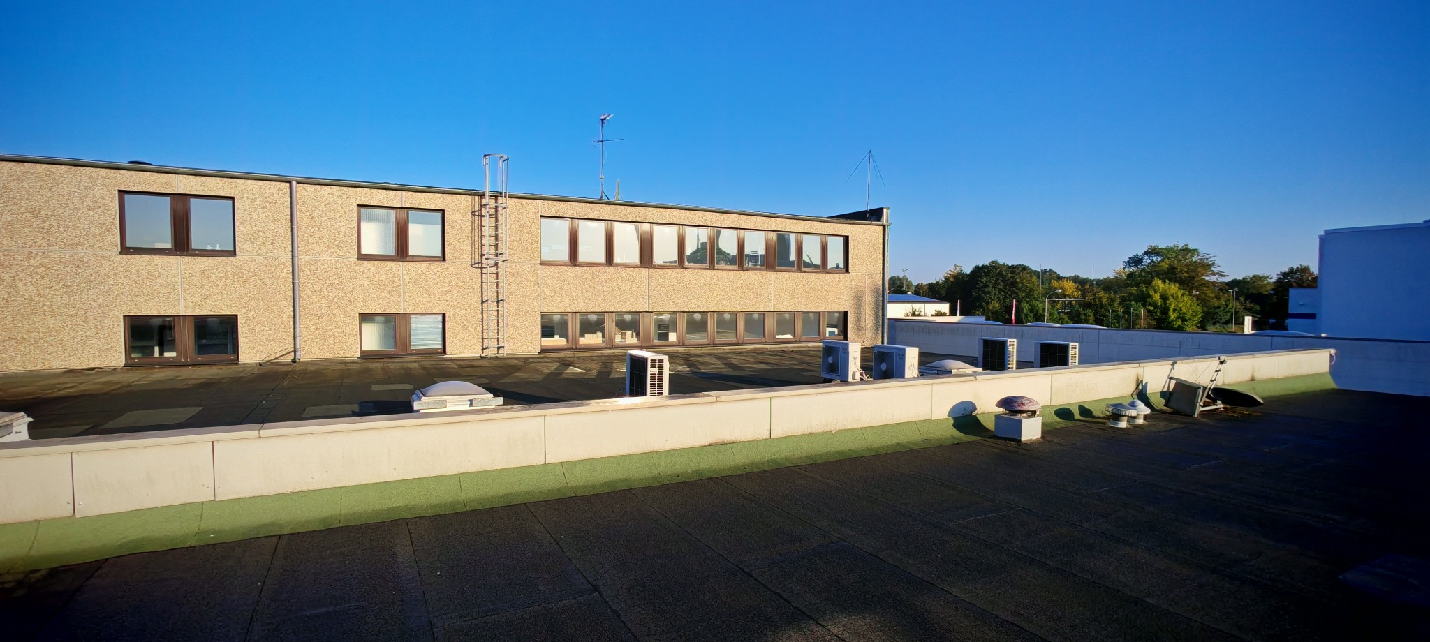 Widok z dachu budynku biurowego na elewację z oknami, drabina ewakuacyjna, anteny i klimatyzatory. Na dachu widoczne świetliki dachowe.