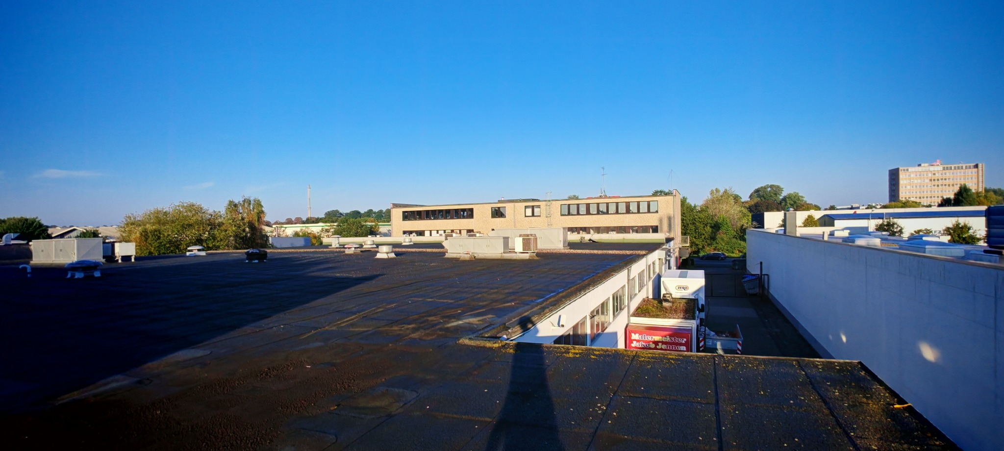 Widok z dachu na budynek biurowy z rzędami okien, anteny i klimatyzatory na dachu, błękitne niebo w tle.