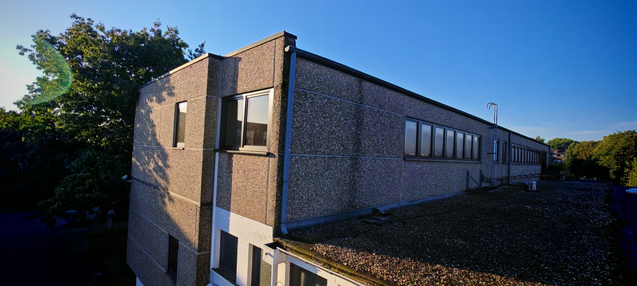 Widok z góry na budynek z oknami w czarnych ramach, elewacja z szarego betonu architektonicznego, na dachu widoczny agregat klimatyzacji i drabina.