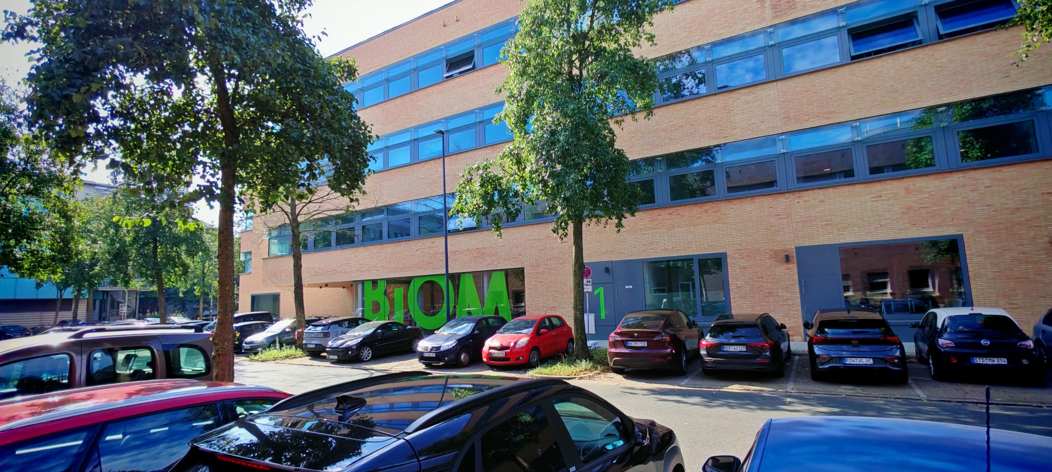 Widok na budynek biurowy z cegły z nowoczesnymi oknami, otoczony zielenią drzew i zaparkowanymi samochodami, z dużym zielonym napisem 'BIOM' nad wejściem.
