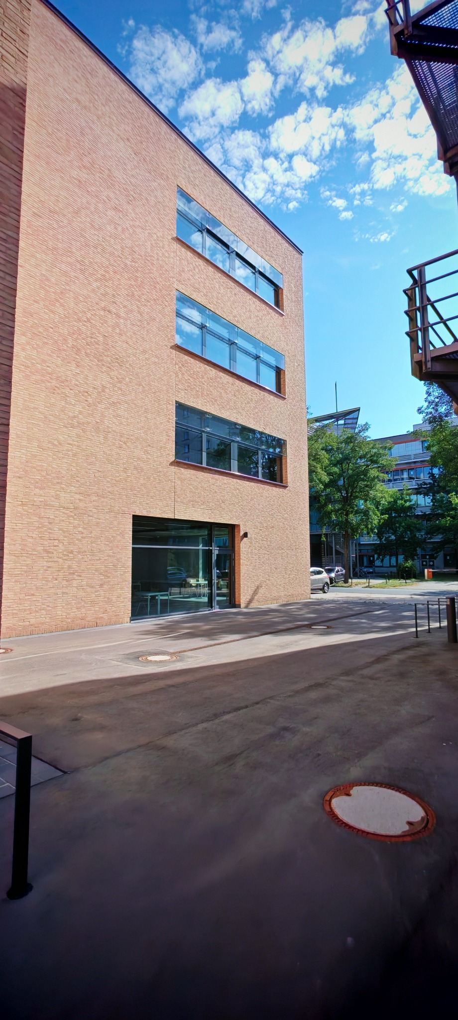 Nowoczesny budynek biurowy z cegły z dużymi, przeszklonymi oknami i wejściem, widziany z poziomu dziedzińca w słoneczny dzień.