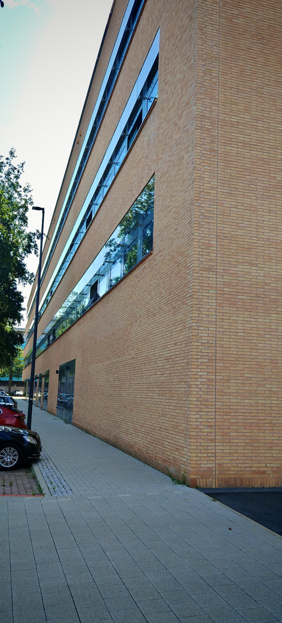 Nowoczesny budynek biurowy z elewacją z jasnej cegły i pasmowymi oknami z ciemnymi ramami, odbijającymi zieleń drzew i błękit nieba.