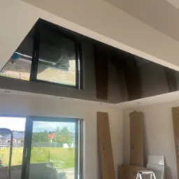 Czarny, lustrzany sufit napinany w nowym domu, odbijający widok przez okno, z widocznymi kartonami i schodkami w tle, sugerujący etap prac wykończeniowych.
