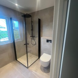 Pasjonaci wnętrz - Nowoczesna łazienka z kabiną prysznicową typu walk-in z czarnymi ramami, wiszącą toaletą i beżowymi płytkami, widok zza uchylonych drzwi.