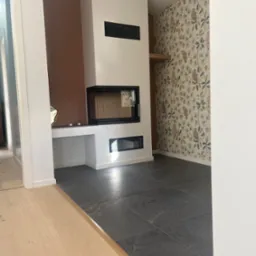 Salon po remoncie z nowoczesnym kominkiem, szarą kamienną podłogą i tapetą w sowy.