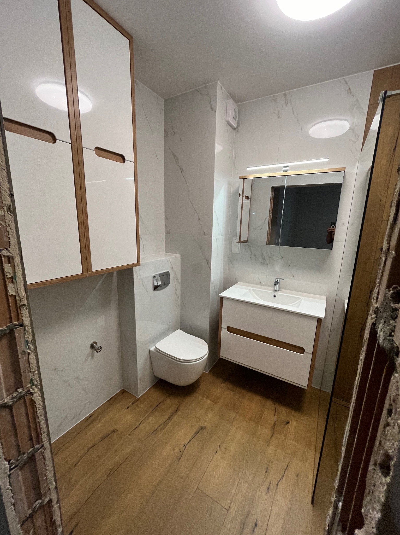 Nowoczesna łazienka po remoncie z drewnopodobną podłogą, białymi szafkami i marmurowymi płytkami ściennymi, widoczne ślady po demontażu ościeżnicy drzwi.