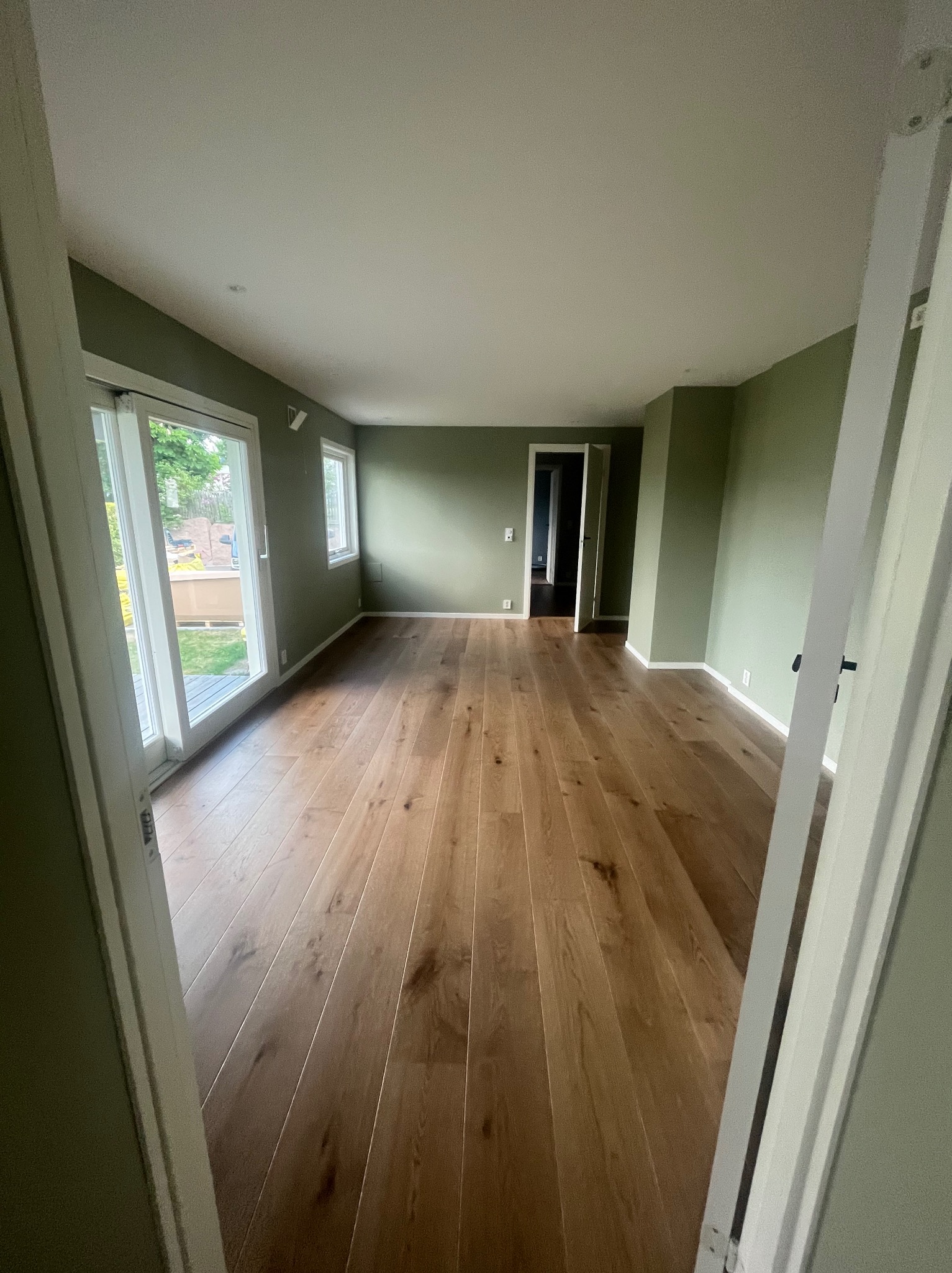 Pusty pokój z drewnianą podłogą w odcieniach brązu, świeżo pomalowanymi ścianami na oliwkowy kolor i białymi listwami przypodłogowymi, widziany z perspektywy otwartych drzwi.