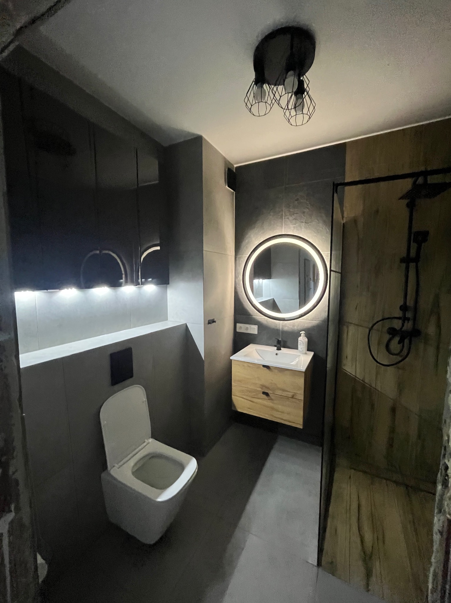 Nowoczesna łazienka z szarymi płytkami, podświetlanym lustrem okrągłym, drewnianą szafką pod umywalką i czarną armaturą prysznicową.