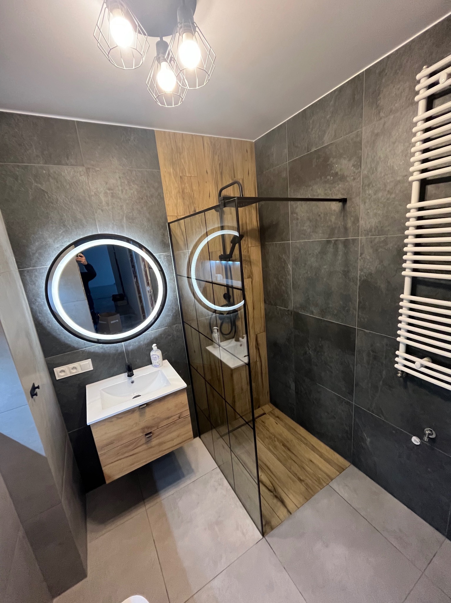 Nowoczesna łazienka z szarymi płytkami, drewnianymi akcentami, okrągłym lustrem z podświetleniem LED, kabiną prysznicową typu walk-in i grzejnikiem drabinkowym.