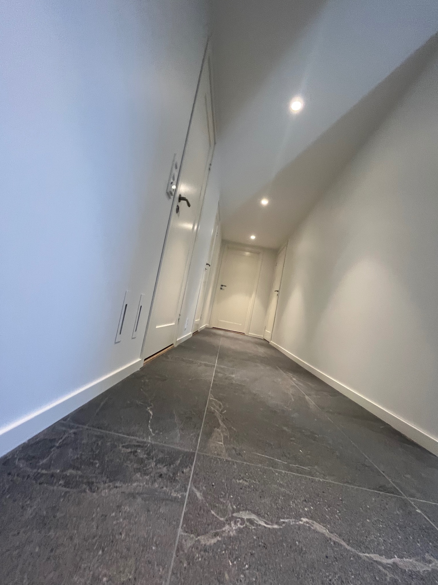 Nowoczesny korytarz z szarymi płytkami imitującymi marmur, białymi ścianami, ukrytymi kratkami wentylacyjnymi i wpuszczanymi lampami sufitowymi.