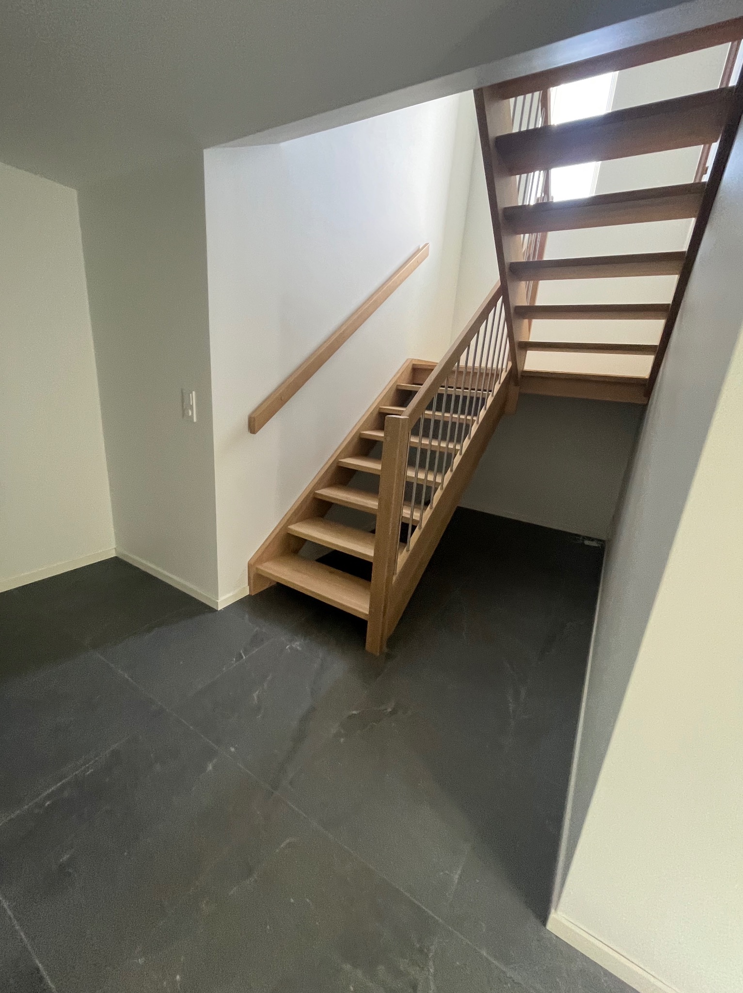 Jasne, drewniane schody z balustradą i poręczą, prowadzące na wyższą kondygnację w nowoczesnym wnętrzu z ciemną, kamienną podłogą.