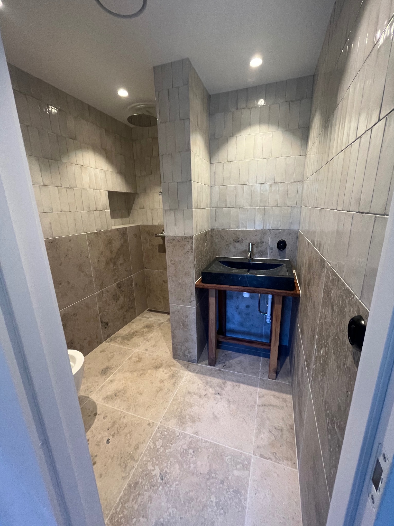 Nowoczesna łazienka z beżowymi płytkami na ścianach i podłodze, czarną umywalką na drewnianym postumencie oraz kabiną prysznicową typu walk-in z deszczownicą.