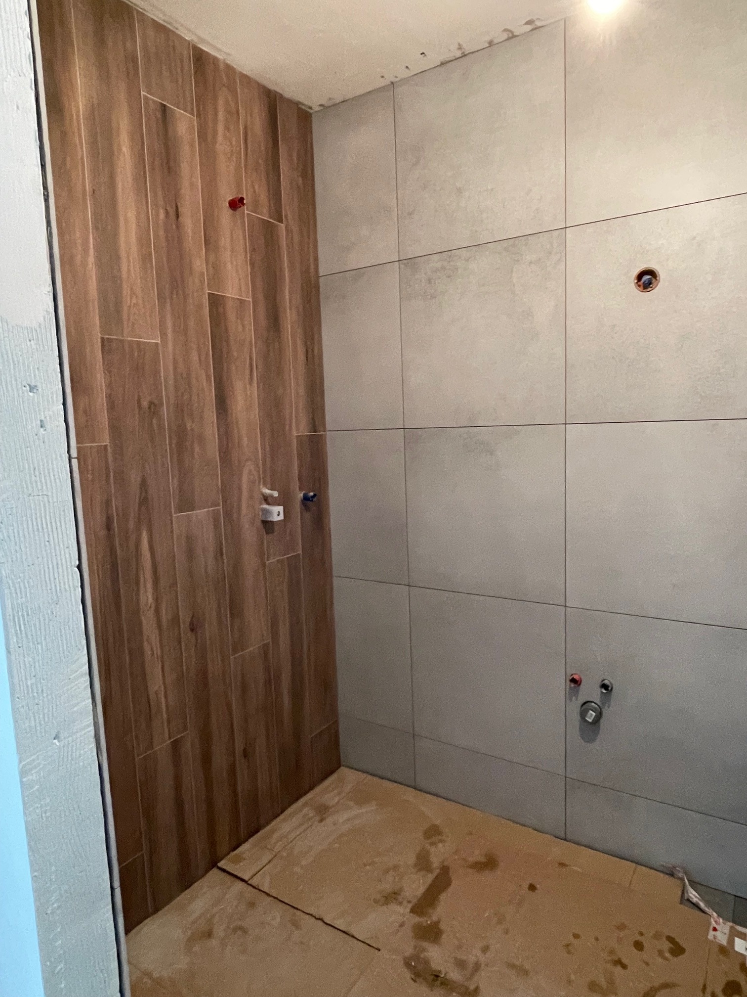 Wnętrze łazienki w trakcie wykańczania: ściany częściowo wyłożone płytkami imitującymi drewno, częściowo szarymi płytkami wielkoformatowymi, zabezpieczona podłoga kartonem, widoczne punkty...