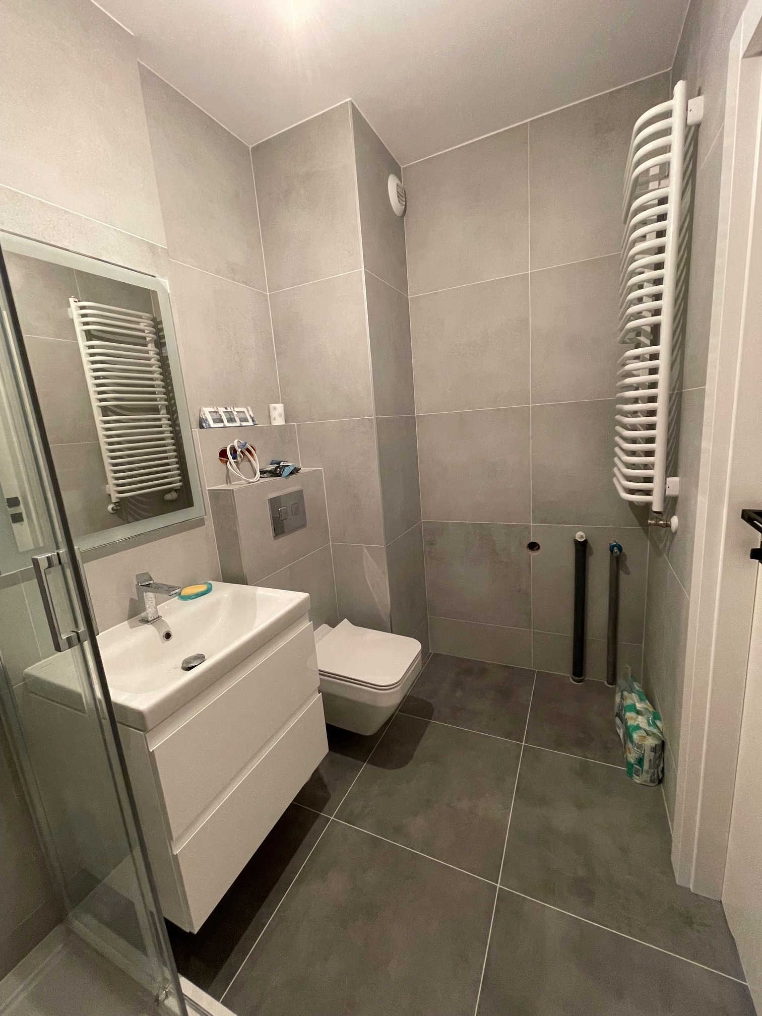 Nowoczesna łazienka w szarościach z białymi elementami: umywalka z szafką, toaleta w zabudowie, grzejnik drabinkowy, szare płytki podłogowe i ścienne, widoczne wystające rury instalacyjne...