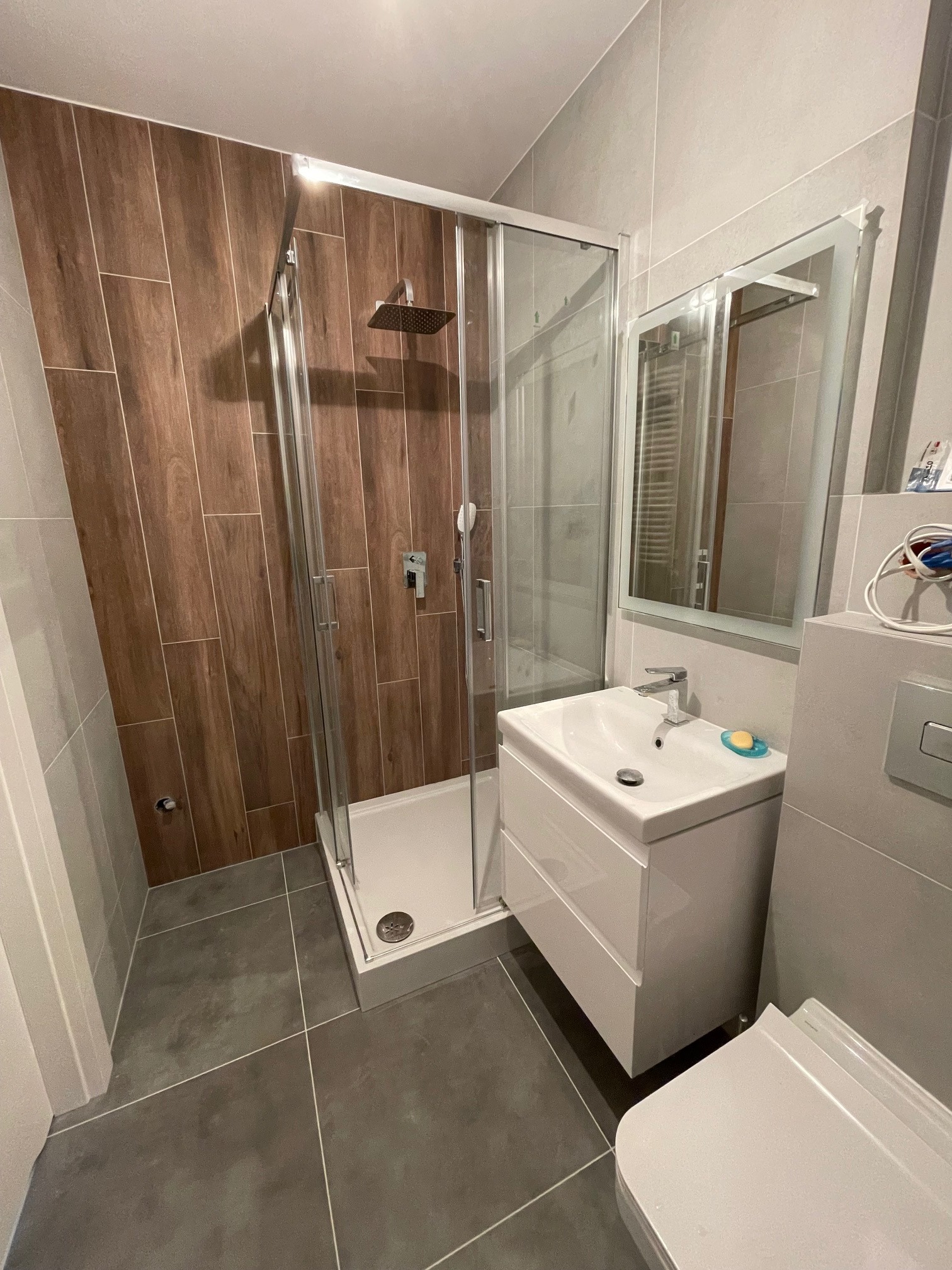 Nowoczesna łazienka po remoncie z kabiną prysznicową, umywalką z szafką, lustrem z podświetleniem LED i płytkami imitującymi drewno na ścianie prysznica.