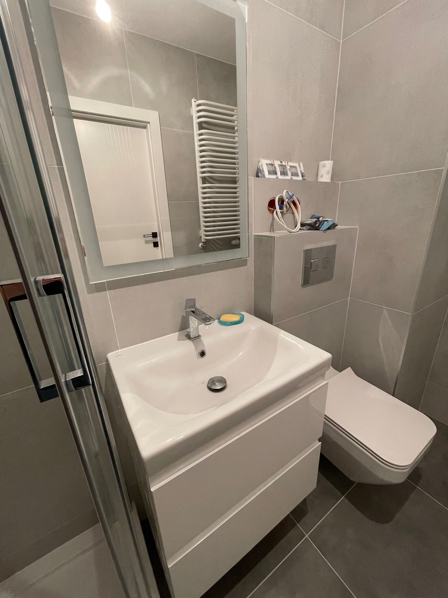 Szara łazienka z umywalką na szafce, toaletą, grzejnikiem drabinkowym i kabiną prysznicową w trakcie wykańczania, widoczne puszki elektryczne i przewody.