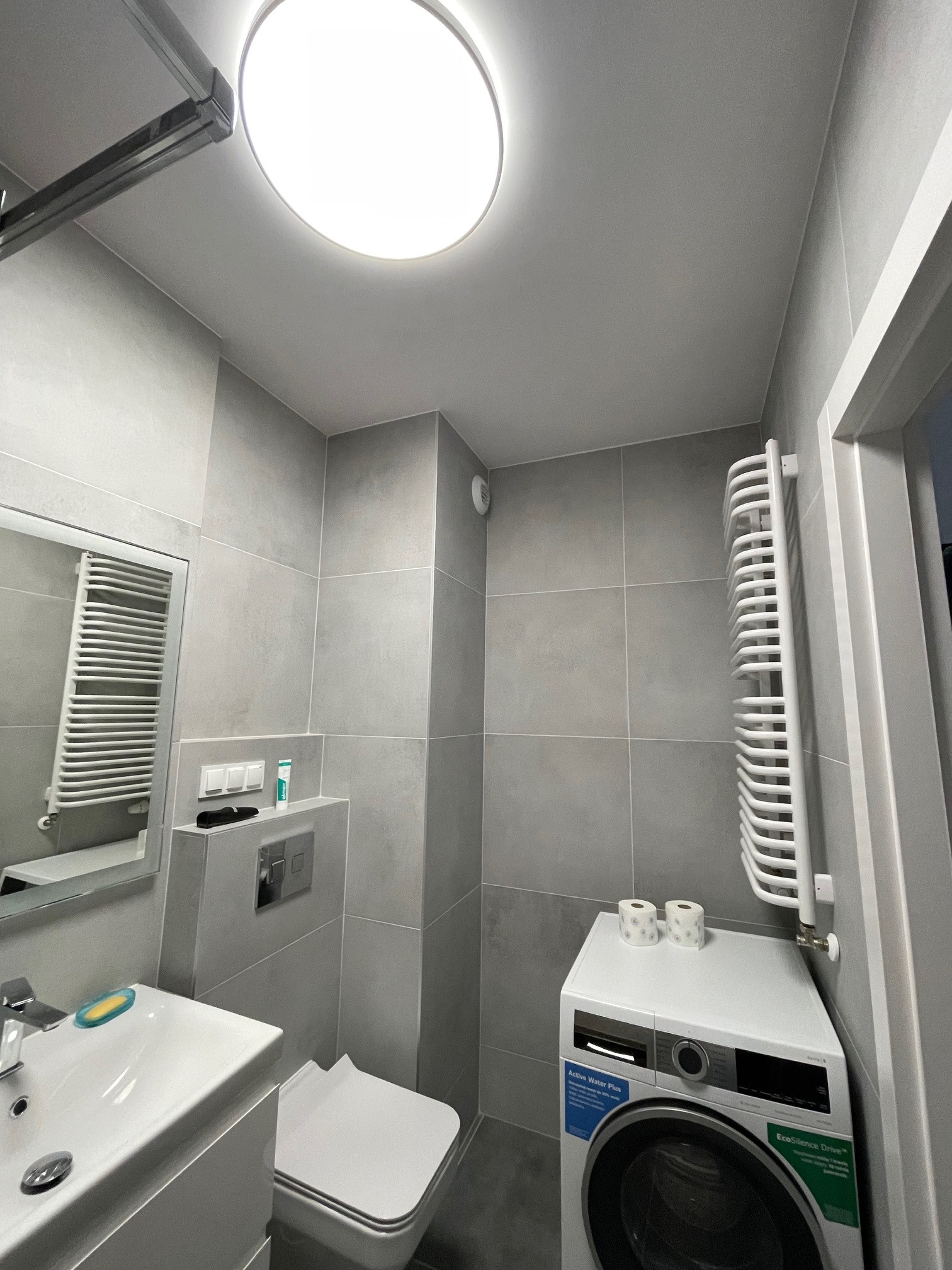 Nowoczesna łazienka w szarościach z płytkami imitującymi kamień, wyposażona w umywalkę z szafką, toaletę, grzejnik drabinkowy, pralkę i okrągłą lampę sufitową.