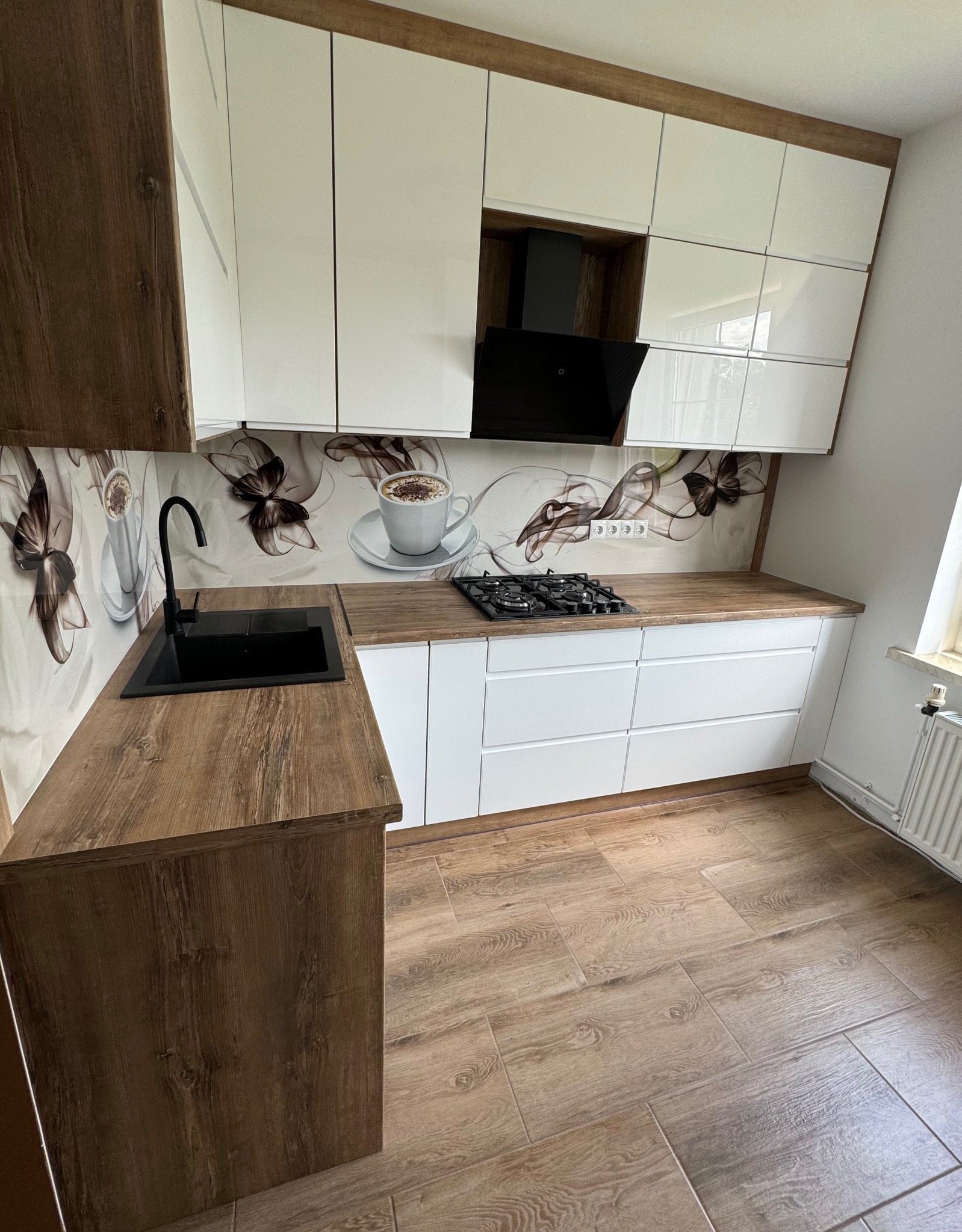 Nowoczesna kuchnia z białymi, lakierowanymi frontami szafek, blatem i wykończeniami w kolorze drewna retro oraz graficznym panelem nad blatem z motywem kawy.